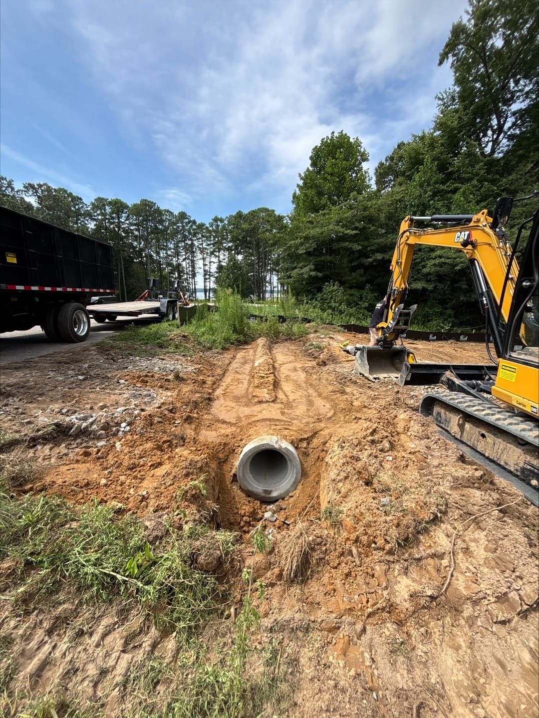 Excavator working on drainage installation with pipe in gravel site under blue sky.
