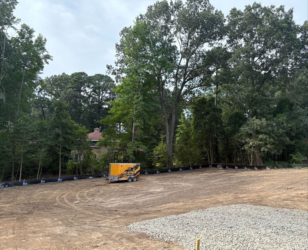 Construction site with a grader and trees in the background. Gravel and silt fence visible.